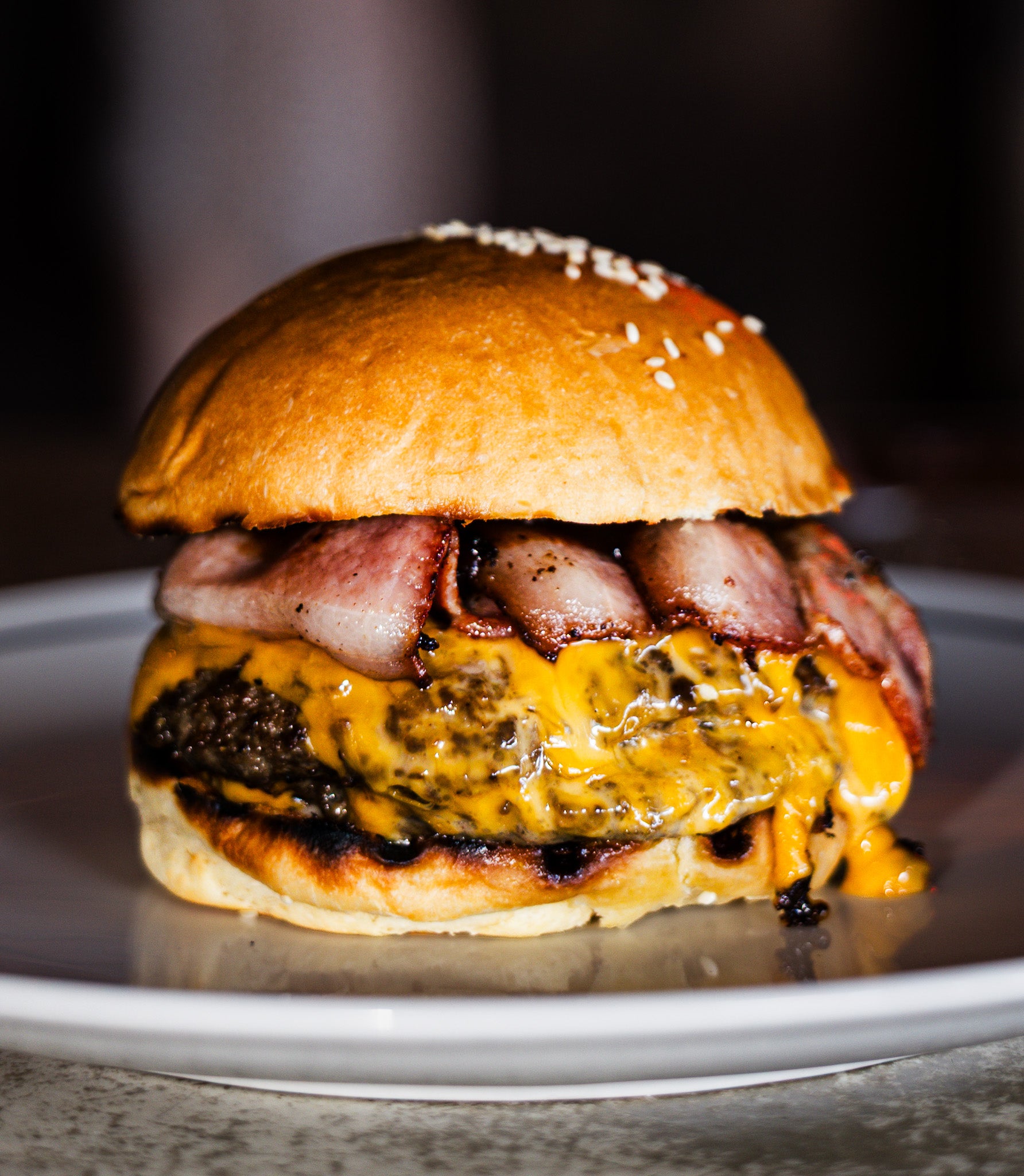 A close-up shot of a gourmet double cheeseburger on a white plate. The burger features a golden-brown sesame seed bun, melted yellow cheese, a grilled beef patty with charred edges, and crispy bacon strips.