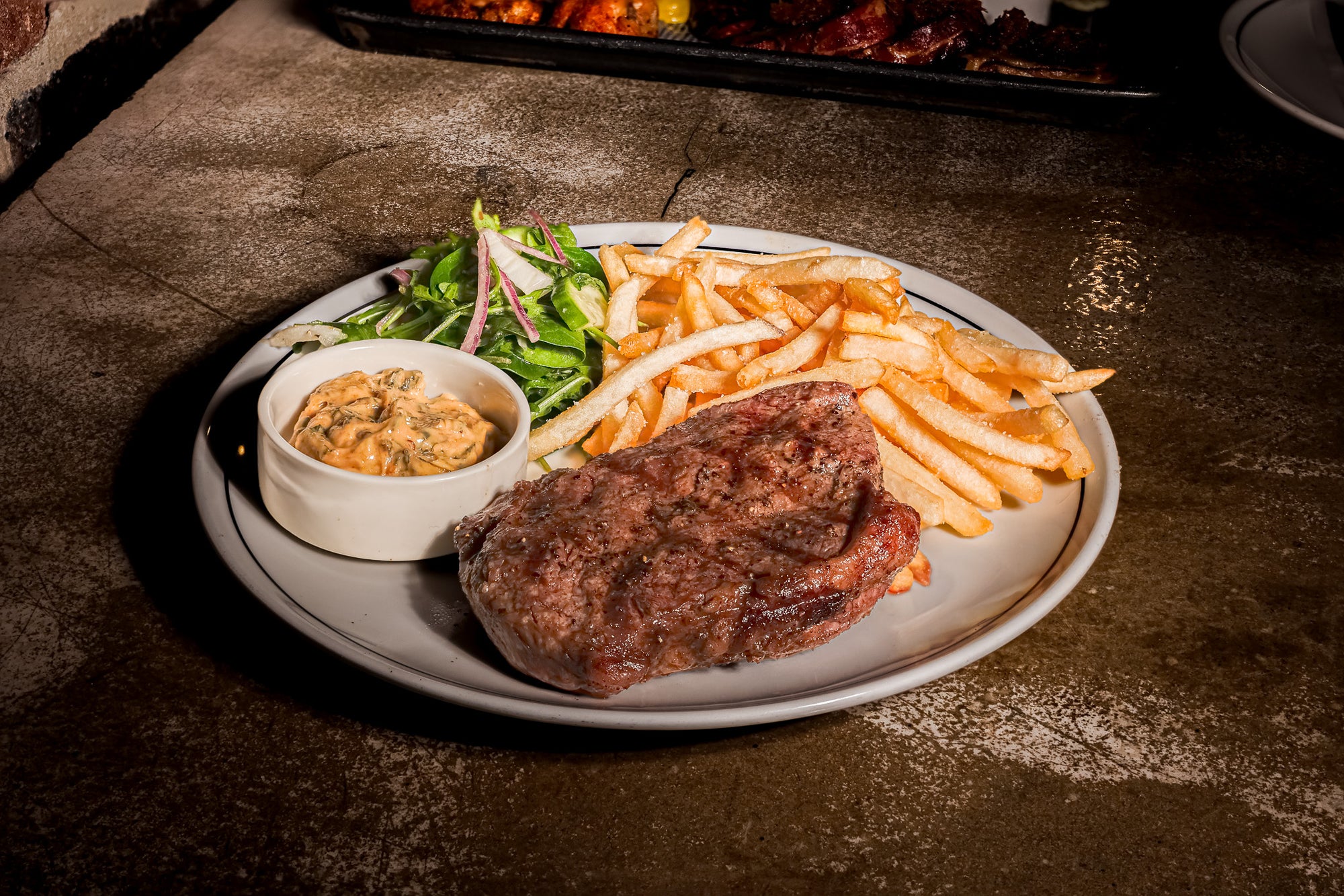 A plated steak with golden fries, a side salad, and a small bowl of cowboy butter on a rustic dark tabletop.
